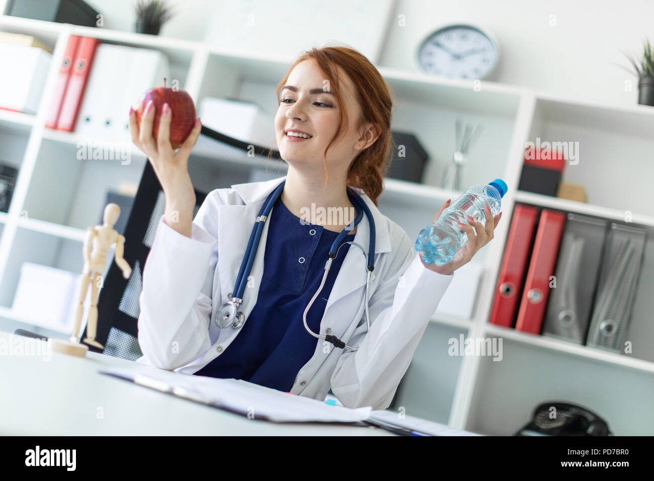 Una hermosa joven vestida con una túnica blanca se sienta en una mesa en la oficina y tiene una manzana y una botella de agua en su mano. Foto de stock