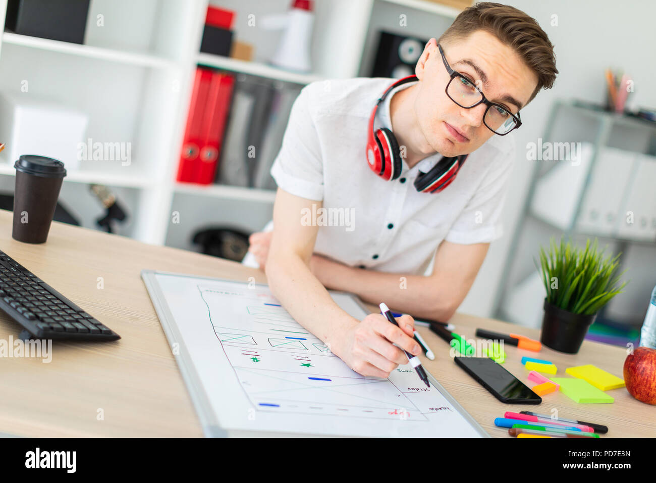 Un hombre joven en copas de pie cerca de una mesa de ordenador. Un hombre joven dibuja un marcador en un tablero magnético. En el cuello, el tío es colgar los auriculares. Foto de stock