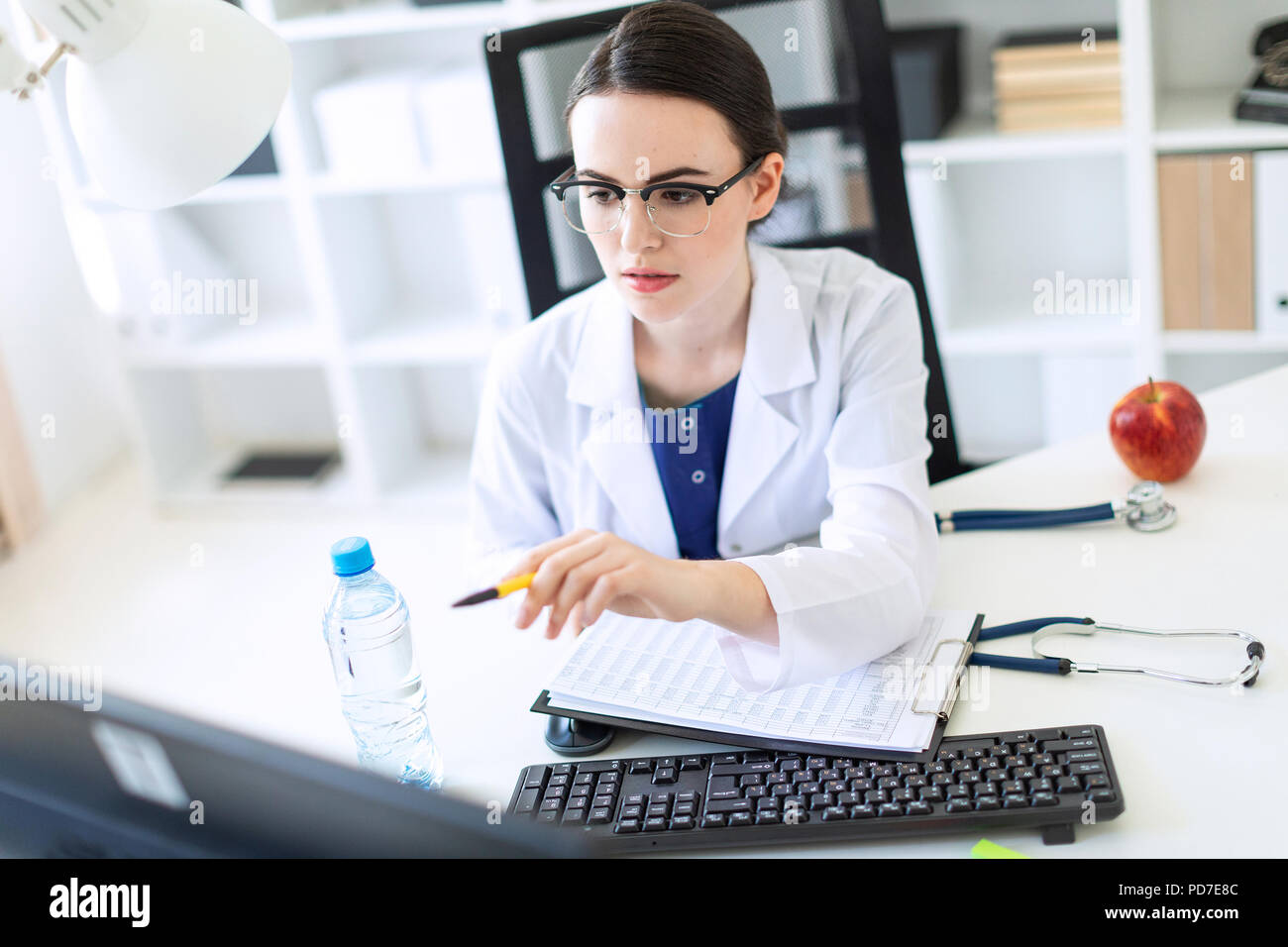 Una hermosa joven vestida con una túnica blanca está sentado frente a una computadora de escritorio con documentos y una pluma en sus manos. Foto de stock