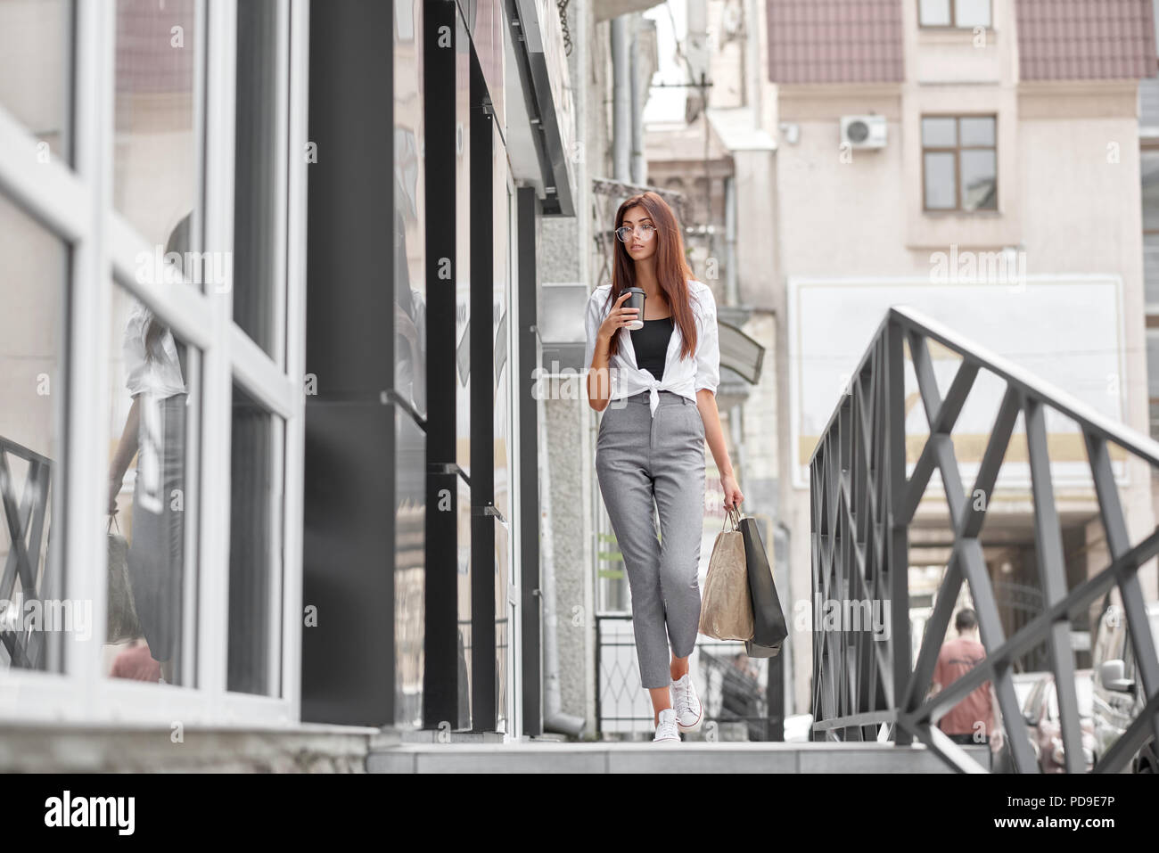 Elegante vestido niña caminando fuera de la tienda de la calle. Llevar  bolsas de papel, manteniendo la taza con café, buscando relajado. Vistiendo  pantalones gris clásico Fotografía de stock - Alamy