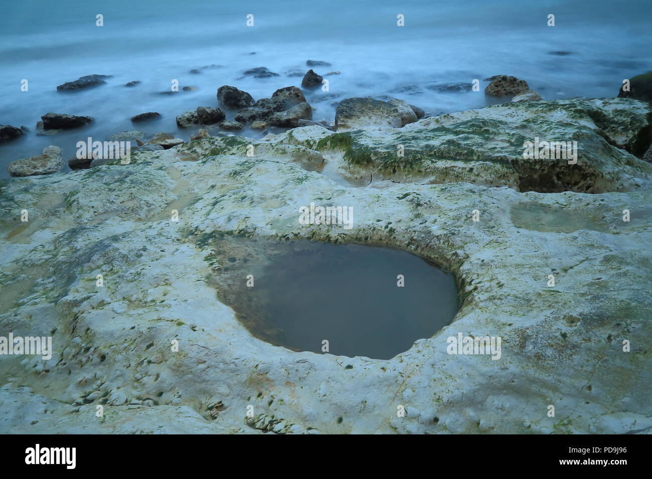 Pequeño estanque salado en la roca caliza sobre la playa de guijarros con olas del mar borrosa Foto de stock