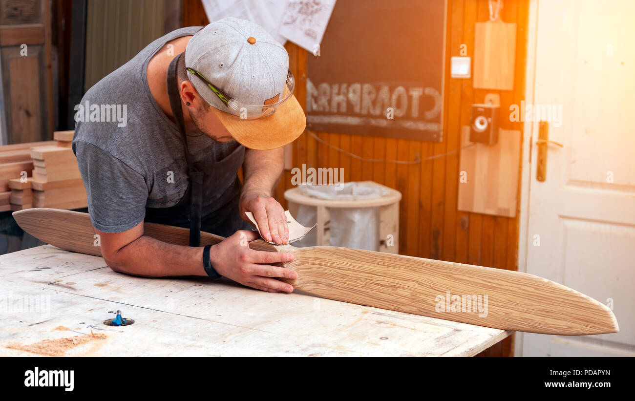 Un hombre de trabajo en una gorra y camisa pule el bloque de madera con  papel de lija antes de pintar en el taller, en el fondo, las herramientas  de perforación y