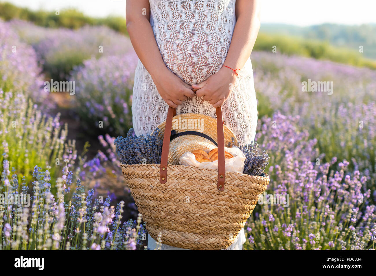 Imagen recortada de la mujer embarazada en vestido blanco celebración cesta de picnic en el campo de lavanda violeta Foto de stock