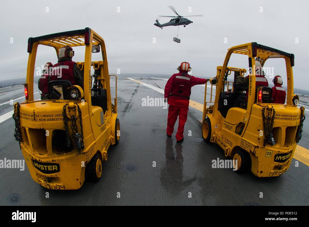 Las aguas al sur de Japón (14 Mayo 2018) departamento de armas marineros de permanecer como un MH-60S Sea Hawk, asignado al comando de Transporte Marítimo Militar (MSC) carga seca/municiones buque USNS César Chávez (T-AKE 14), ofrece detonar en la cubierta de vuelo a bordo de la Marina desplegadas, el portaviones USS Ronald Reagan (CVN 76), como parte de una reposición en alta mar durante las pruebas de mar. Los civiles no combatientes, con tripulación de barco, operado por la MSC, proporciona el combustible, alimentos, municiones, piezas de repuesto, el correo y otros suministros a los buques de la Marina en todo el mundo. Ronald Reagan, el buque insignia de Carrier Strike Group 5, proporciona Foto de stock