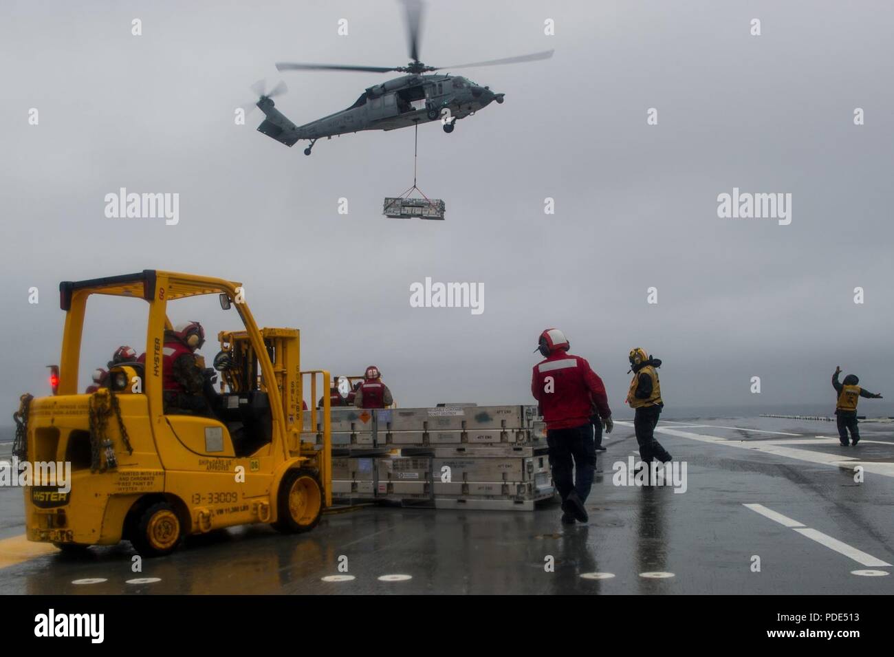 Las aguas al sur de Japón (14 Mayo 2018) departamento de armas marineros de permanecer como un MH-60S Sea Hawk, asignado al comando de Transporte Marítimo Militar (MSC) carga seca/municiones buque USNS César Chávez (T-AKE 14), ofrece detonar en la cubierta de vuelo a bordo de la Marina desplegadas, el portaviones USS Ronald Reagan (CVN 76), como parte de una reposición en alta mar durante las pruebas de mar. Los civiles no combatientes, con tripulación de barco, operado por la MSC, proporciona el combustible, alimentos, municiones, piezas de repuesto, el correo y otros suministros a los buques de la Marina en todo el mundo. Ronald Reagan, el buque insignia de Carrier Strike Group 5, proporciona Foto de stock