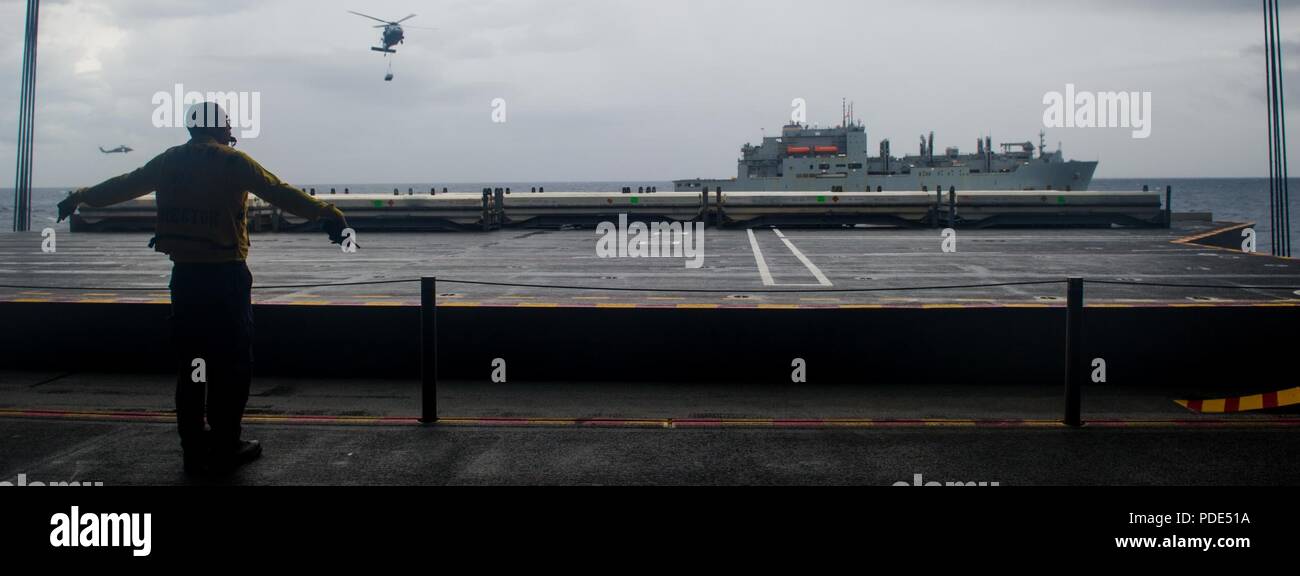 Las aguas al sur de Japón (14 de mayo de 2018) la aviación Boatswain's Mate (manejo) de primera clase Rashad Gloster indica el descenso de una aeronave elevador como MH-60S mar, halcones, asignado al comando de Transporte Marítimo Militar (MSC) carga seca/municiones buque USNS César Chávez (T-AKE 14), entregar explosivos a la Marina desplegadas, el portaviones USS Ronald Reagan (CVN 76), como parte de una reposición en alta mar durante las pruebas de mar. Los civiles no combatientes, con tripulación de barco, operado por la MSC, proporciona el combustible, alimentos, municiones, piezas de repuesto, el correo y otros suministros a los buques de la Marina en todo el mundo. Ronald Reagan, el buque insignia Foto de stock