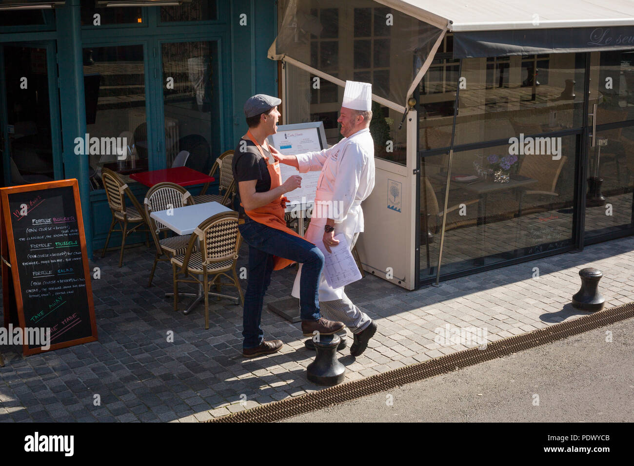 Un chef disfruta de una broma con un amigo en una escena callejera en Honfleur, Normandía, Francia Foto de stock