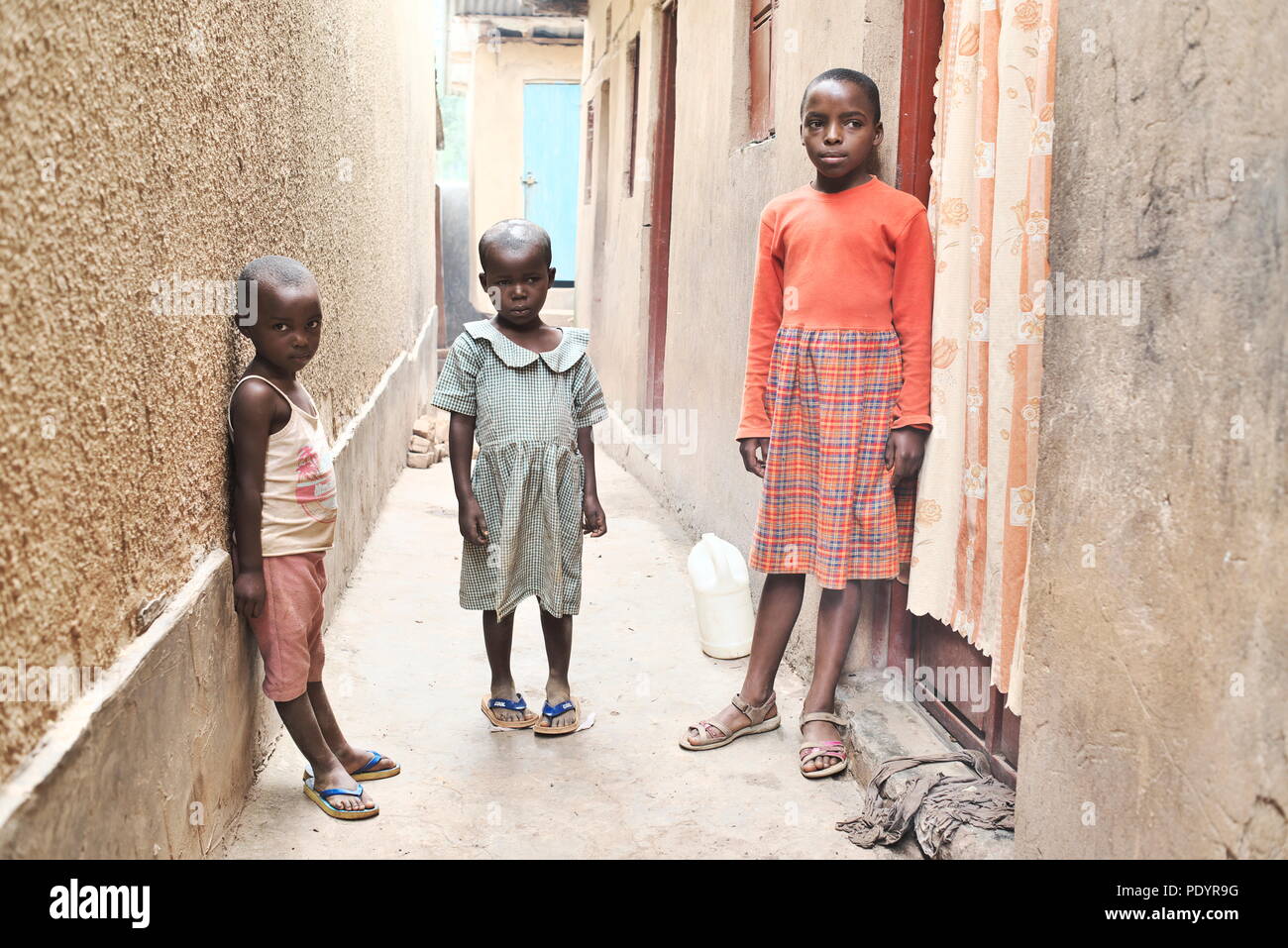 Tres niños africanos jóvenes pobres se encuentran en un callejón en Uganda, en un barrio marginal, vestidos con ropa básica fuera de sus hogares pobres Foto de stock