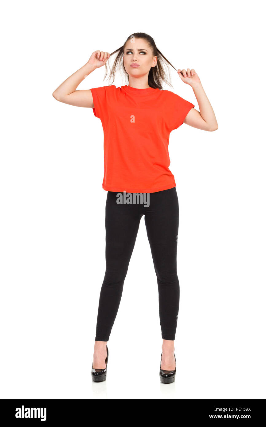 Muecas joven con leggings negro, tacones altos y camiseta naranja está de pie, sosteniendo su cabello y apartar la mirada. Foto de estudio aislado de longitud completa Foto de stock