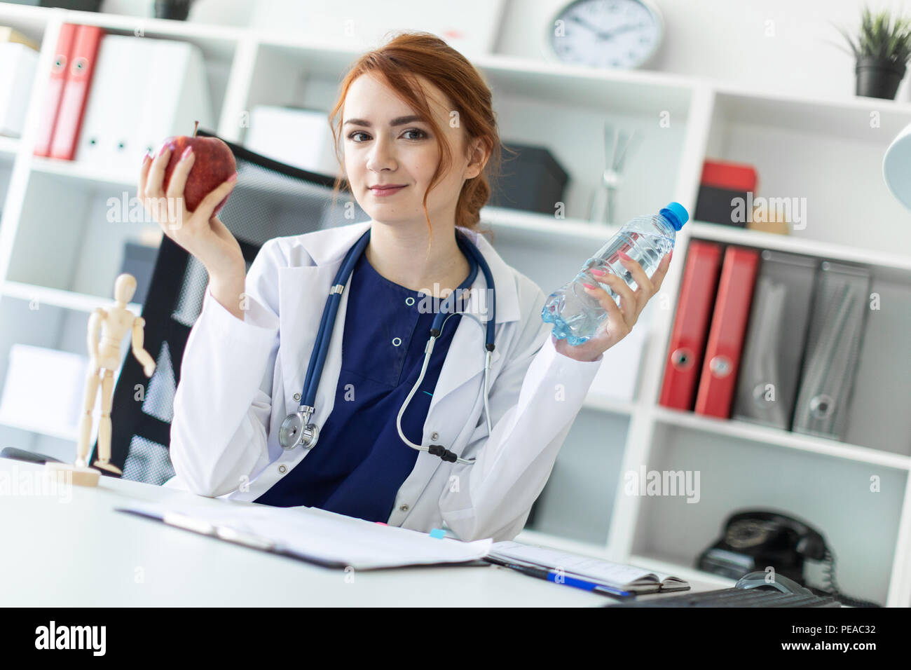 Una hermosa joven vestida con una túnica blanca se sienta en una mesa en la oficina y tiene una manzana y una botella de agua en su mano. Foto de stock