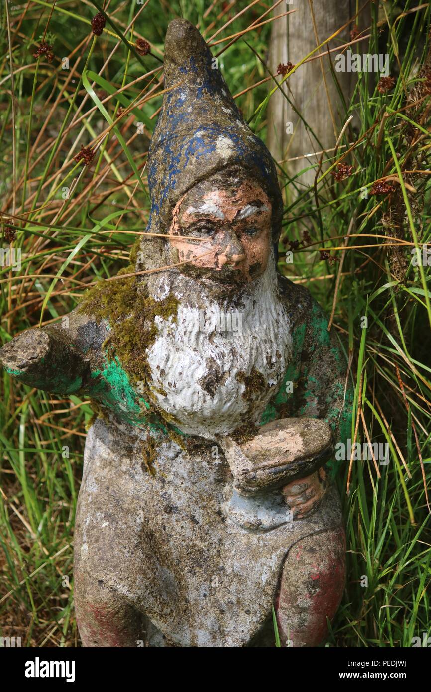 Muy viejo y tiempo gastado gnomo de jardín con barba blanca, chaqueta cubiertas de musgo y barro tapa sombrero puntiagudo holding y yunque y martillo Foto de stock