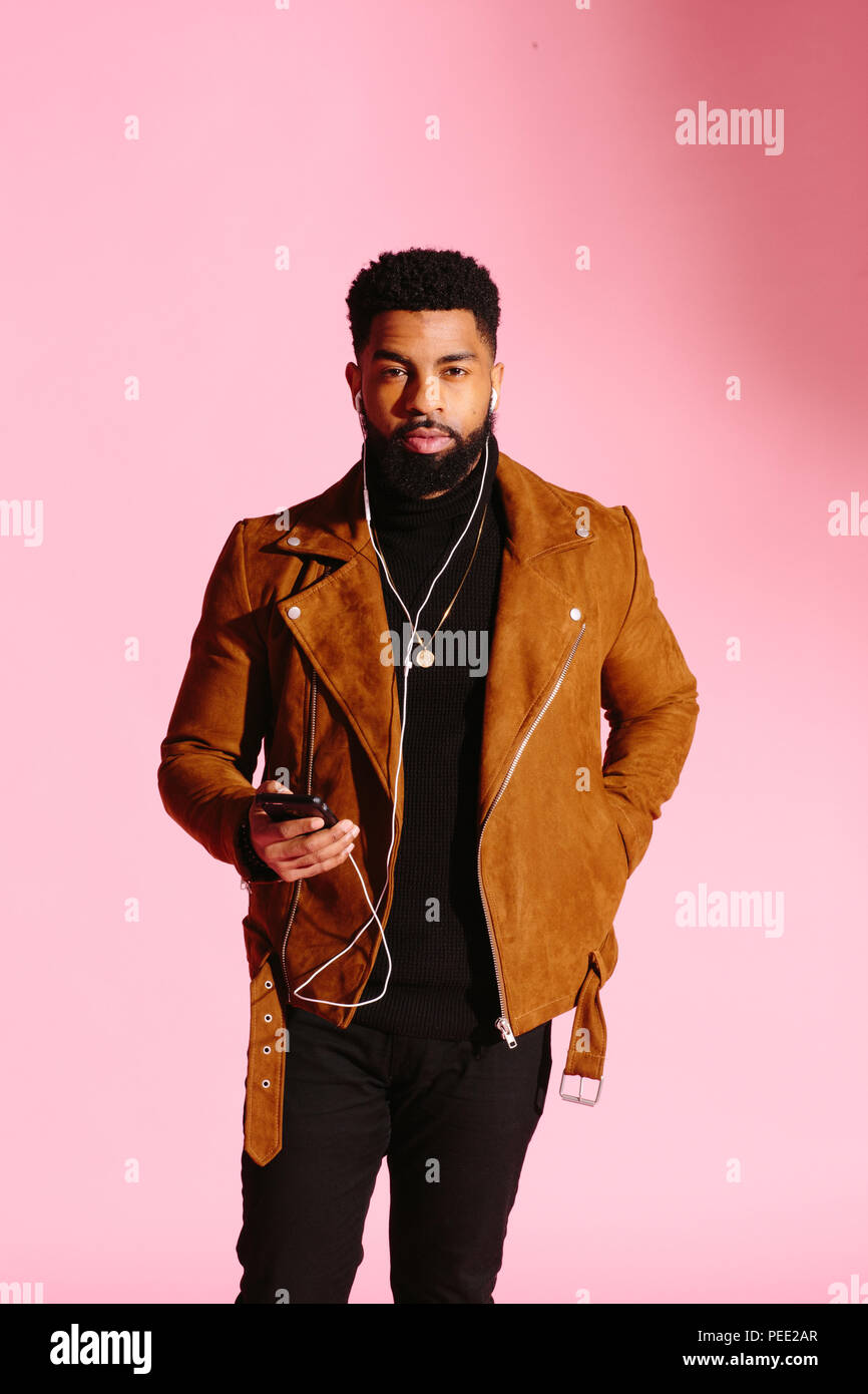 Elegante, guapo y cool hombre afroamericano con barba, escuchando música, mensajes de texto, aislado sobre fondo rosa studio Foto de stock