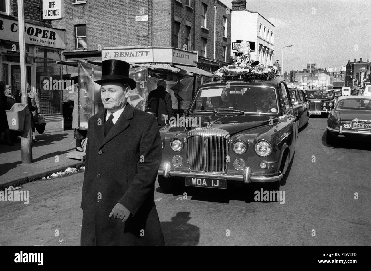 Funeral de Londres de 1970. Hoxton director funeral oficial en su top hat camina en frente carroza automóviles siga en procesión Hoxton Street 70s Inglaterra HOMER SYKES. Foto de stock