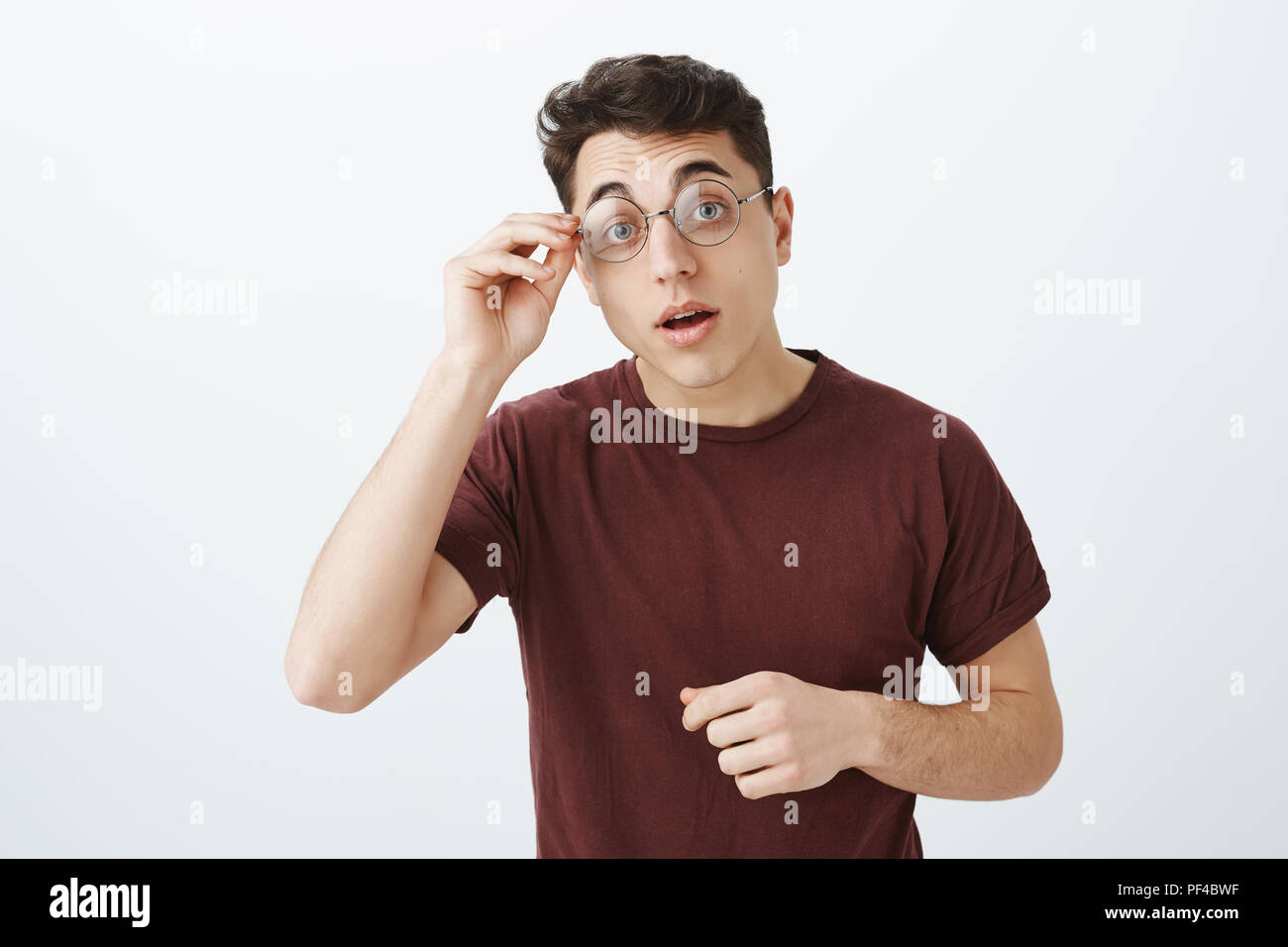 Foto de estudio de smart curioso guapo en gafas redondas y camiseta roja,  levantamiento de cejas con interés y sosteniendo el aro de gafas ver caso  interesante, querer resolver enigma o asignación