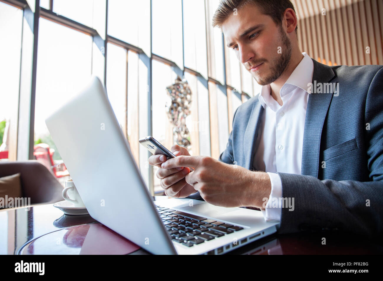 Empresario de traje negro con mobile teléfono inteligente y trabajar en equipo portátil, navegar por internet y escribir en el bloc de notas de papel en la oficina moderna. Hombre trabajando en dispositivos electrónicos con espacio de copia Foto de stock