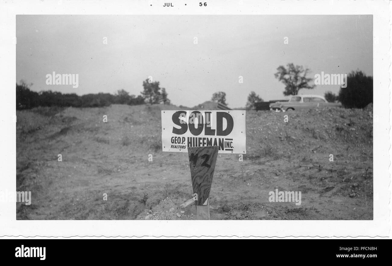Fotografía en blanco y negro, mostrando un cartel con las palabras "viejo, GEO P Realtors, Huffman Inc" en un sitio de construcción con montículos de tierra en primer plano y un automóvil vintage visibles en el fondo, probablemente fotografiado en Ohio, julio de 1956. () Foto de stock