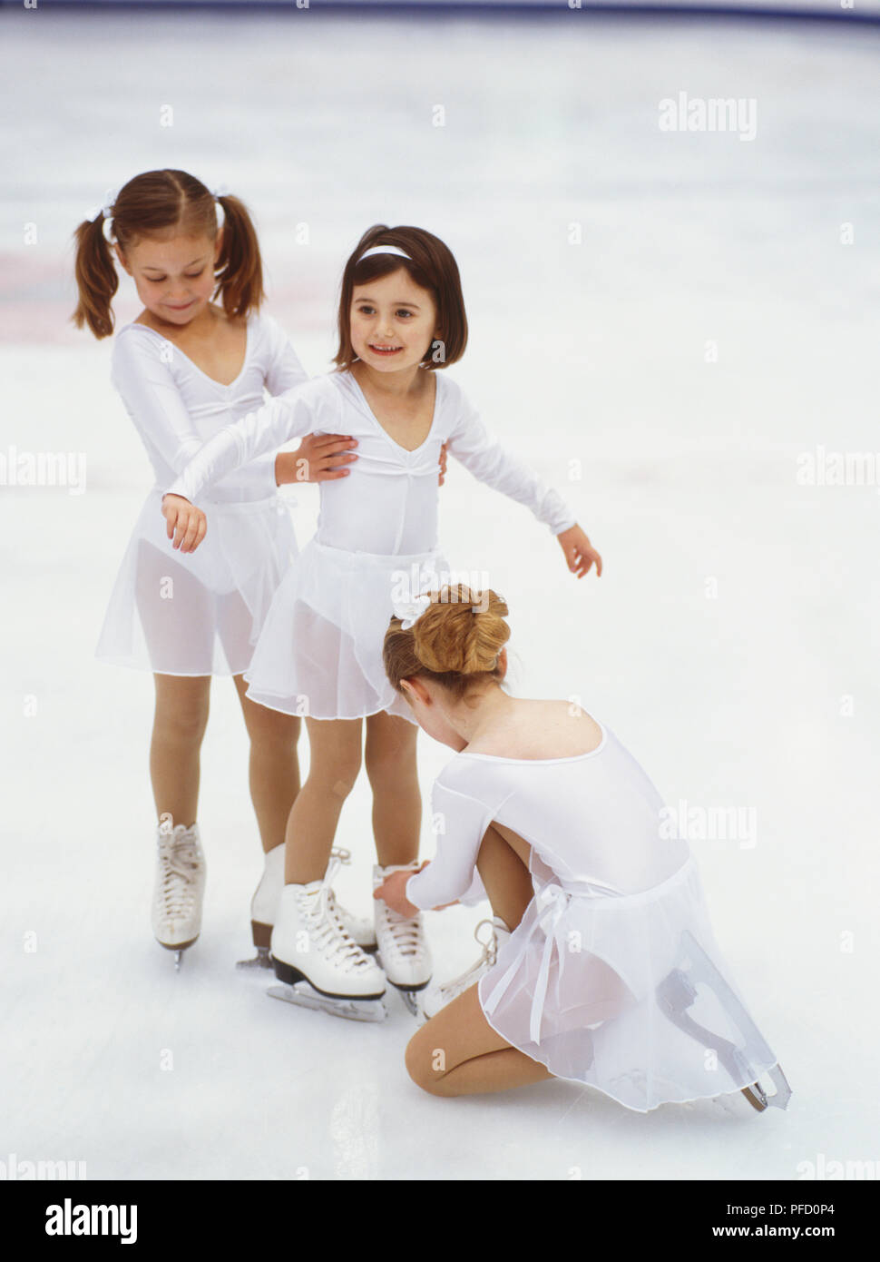 Grupo de tres niños en blanco trajes de patinaje sobre hielo, primera niña  apoyar una segunda, tercera niña arrodillarse sobre hielo y atar los  cordones de zapatos de la segunda chica Fotografía