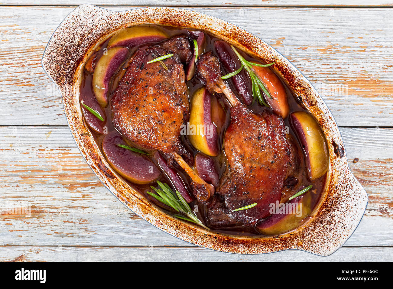 Las piernas de pato asado en horno cocido en vino tinto con romero,  especias y rodajas de manzana en la bandeja de hornear, sobre blanco  pintura desconchada rústico boards, vista desde ab