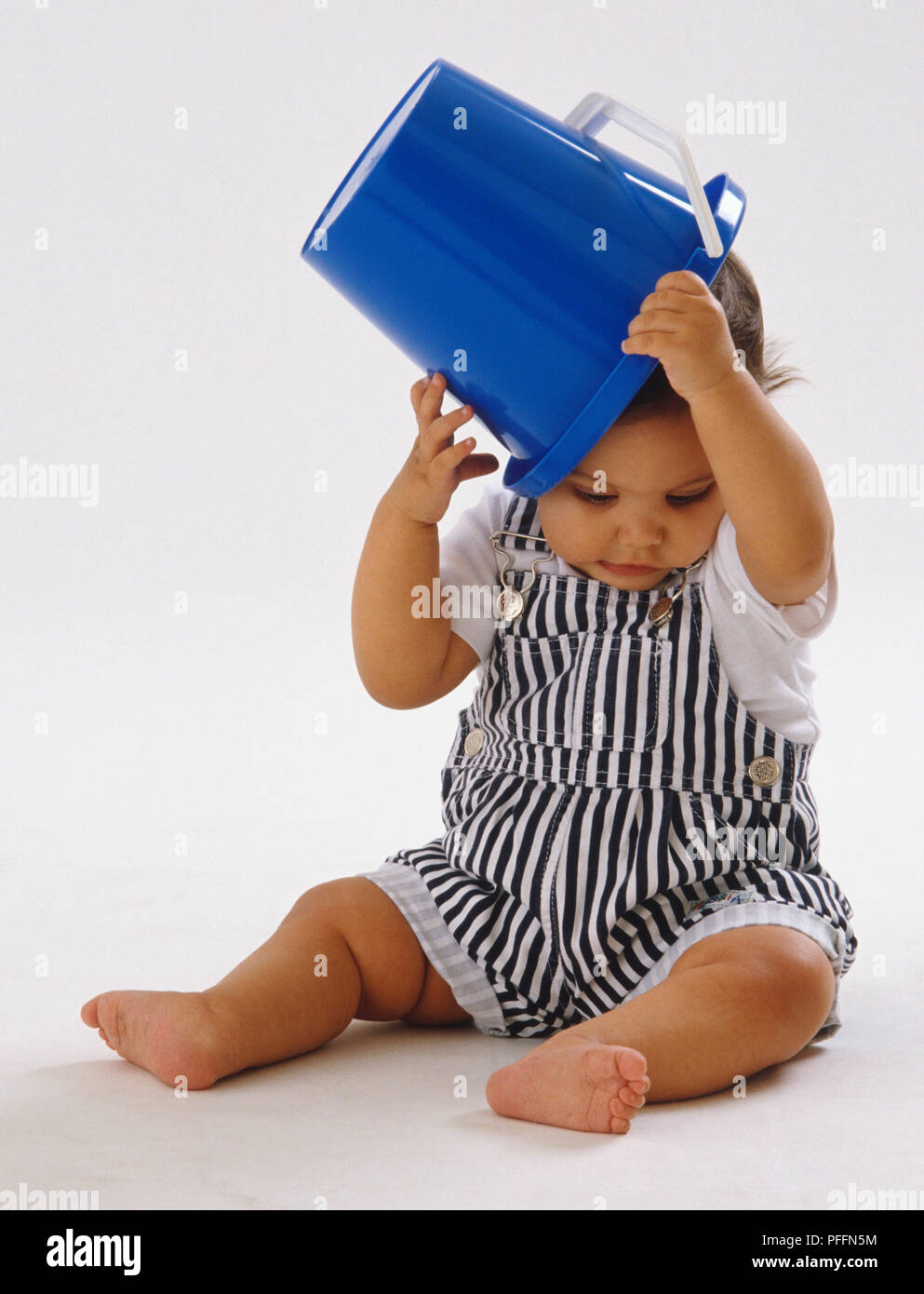 Bebé vistiendo petos rayados, sentarse, poner pucherito cuchara azul encima de su cabeza. Foto de stock