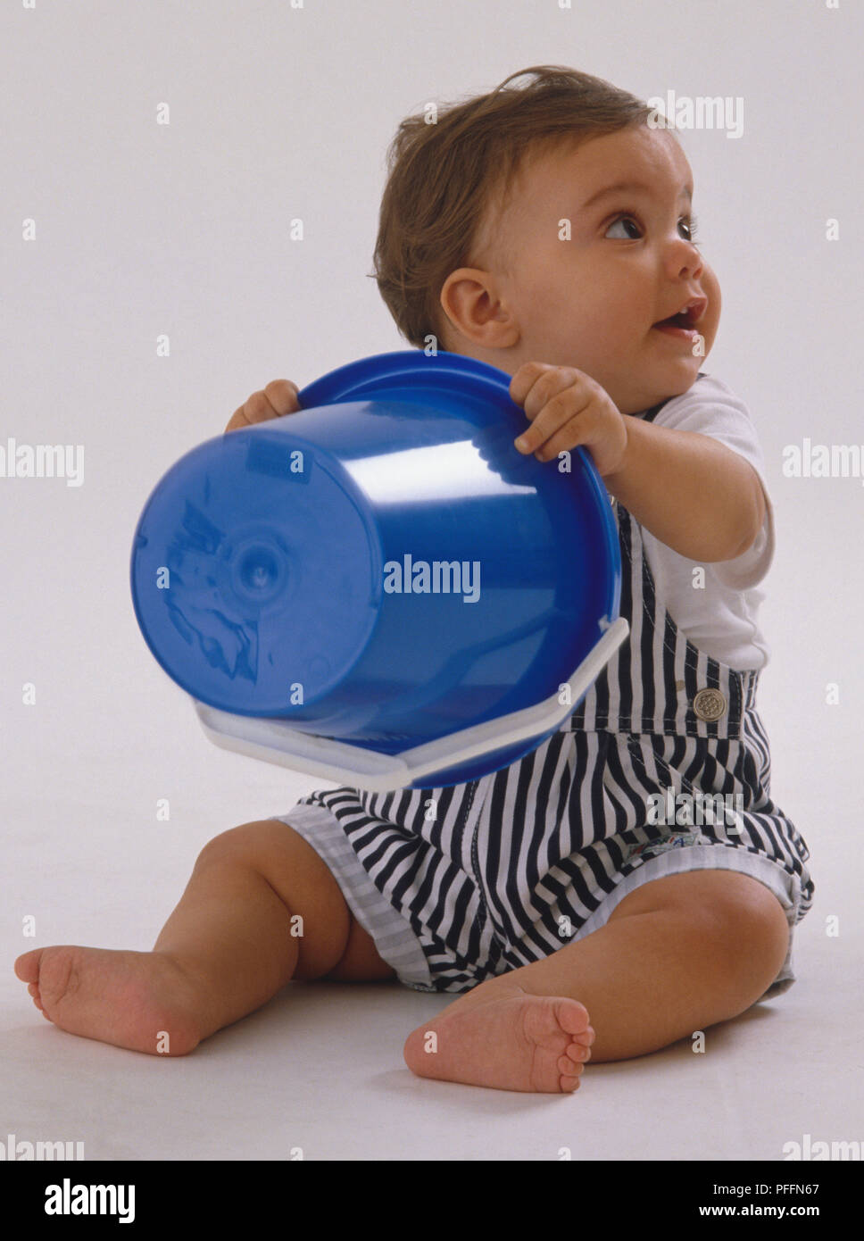 Bebé vistiendo petos rayados, agarrando la cuchara de plástico azul, mirando hacia arriba, sonriendo. Foto de stock