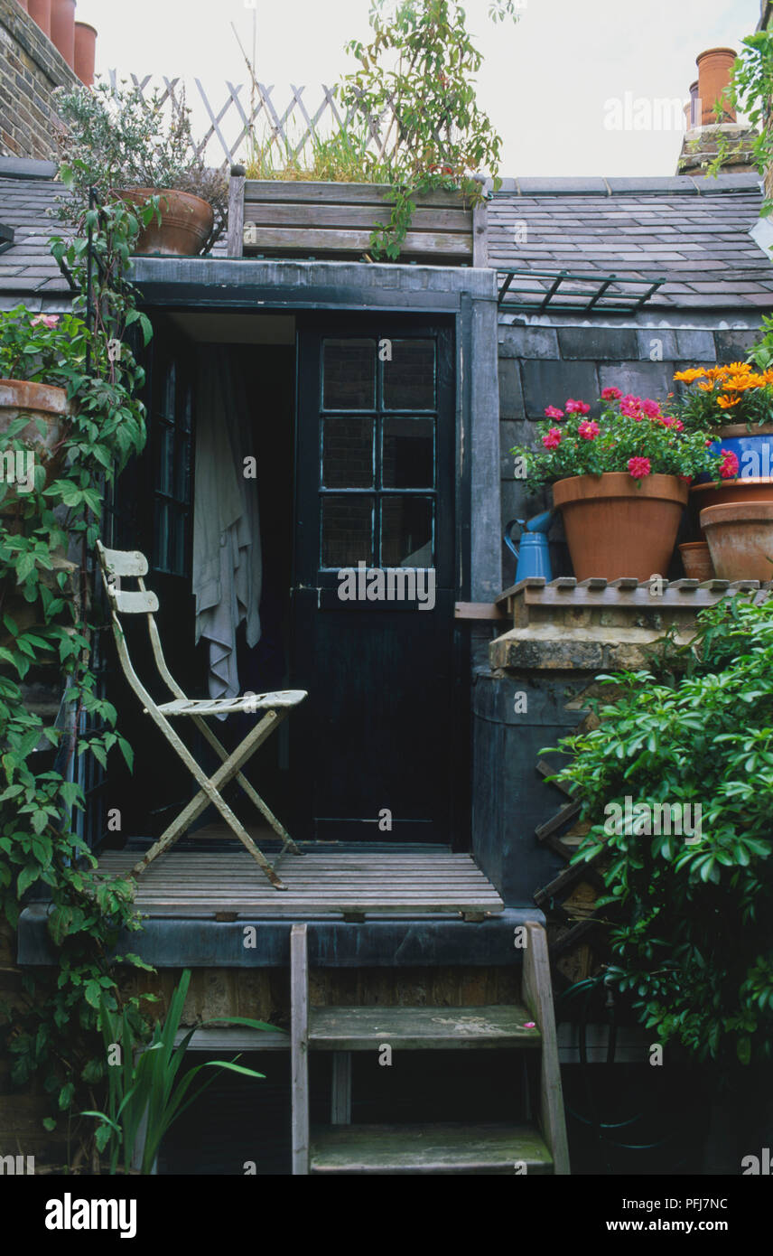 Terraza pequeña con la vieja silla, verde oscuro, con doble puerta semiabierta, toallas grandes colgando sobre la puerta, enmarcado por viñedos y macetas, bajo cubierta de teja negra Foto de stock