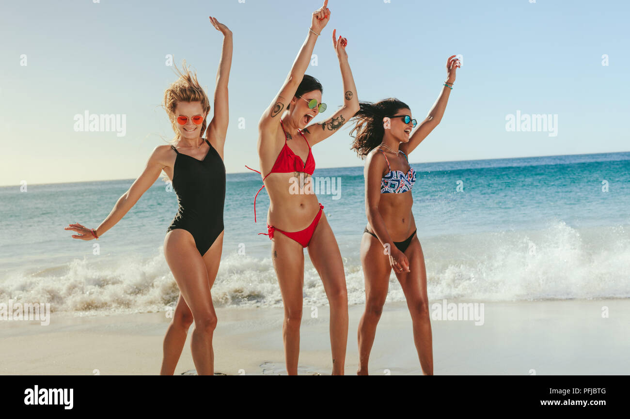 Tres amigas en bikini con gafas de sol bailando en la playa para levantar  los brazos. La mujer feliz de vacaciones tomando el sol y disfrutando en la  playa con vistas al