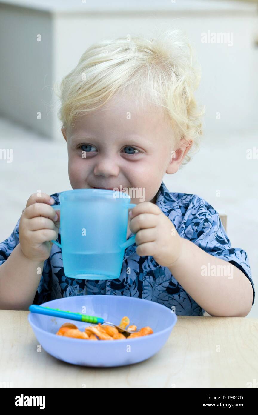 Baby Boy rubia sosteniendo un vaso de plástico sobre el tazón de comida, close-up Foto de stock