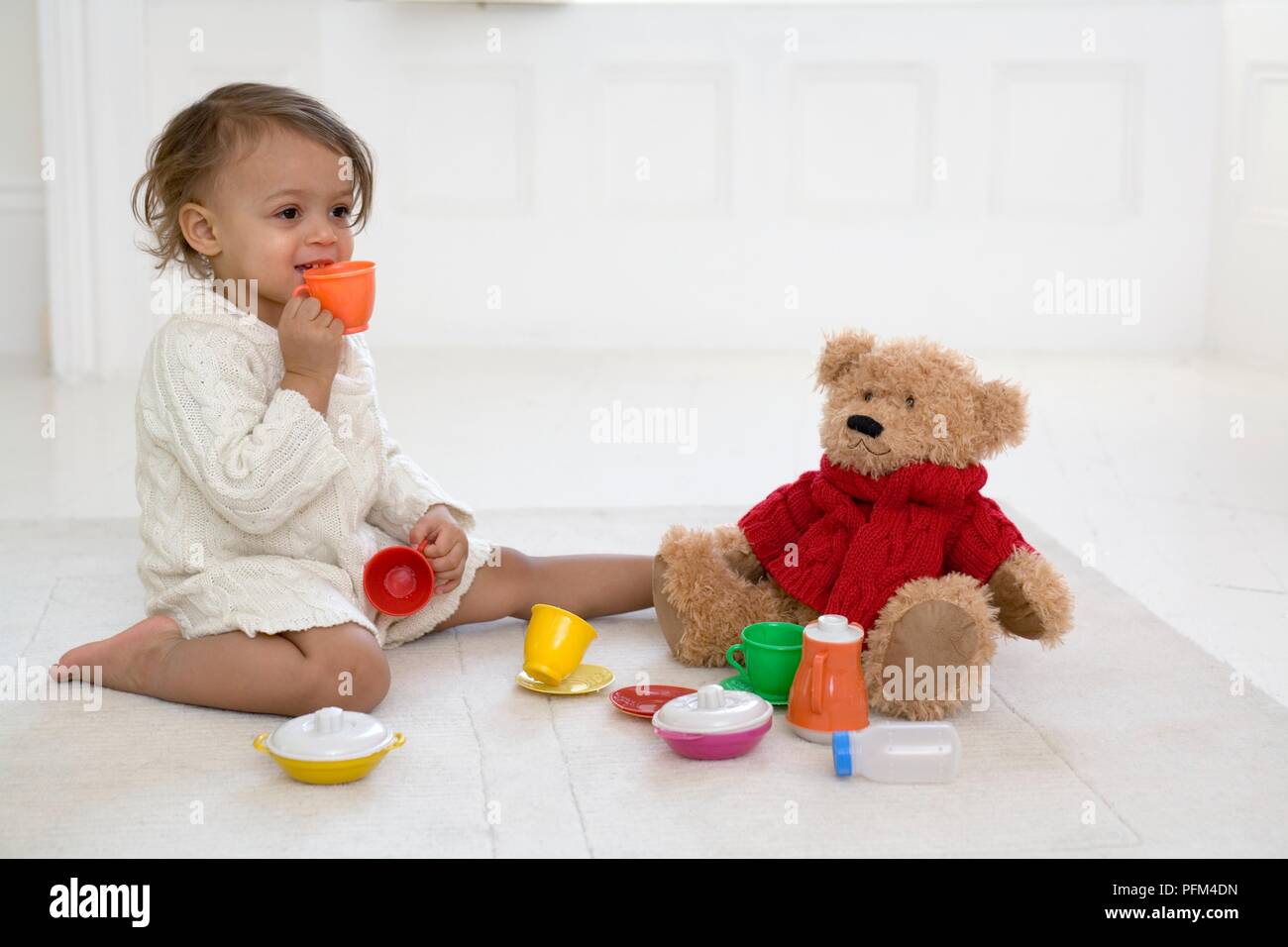 Chica sentada en el suelo sujetando un vaso de plástico a su boca, osito de peluche y diversos artículos de plástico a su alrededor. Foto de stock