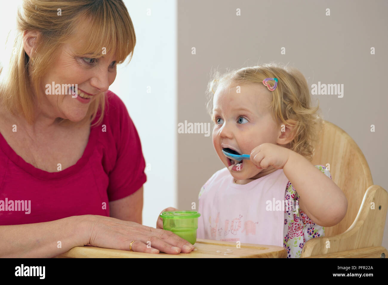 Abuela sentada junto a su nieta en la silla alta y la celebración de la olla, mientras que su nieta se alimenta a sí misma desde una cuchara de plástico azul Foto de stock