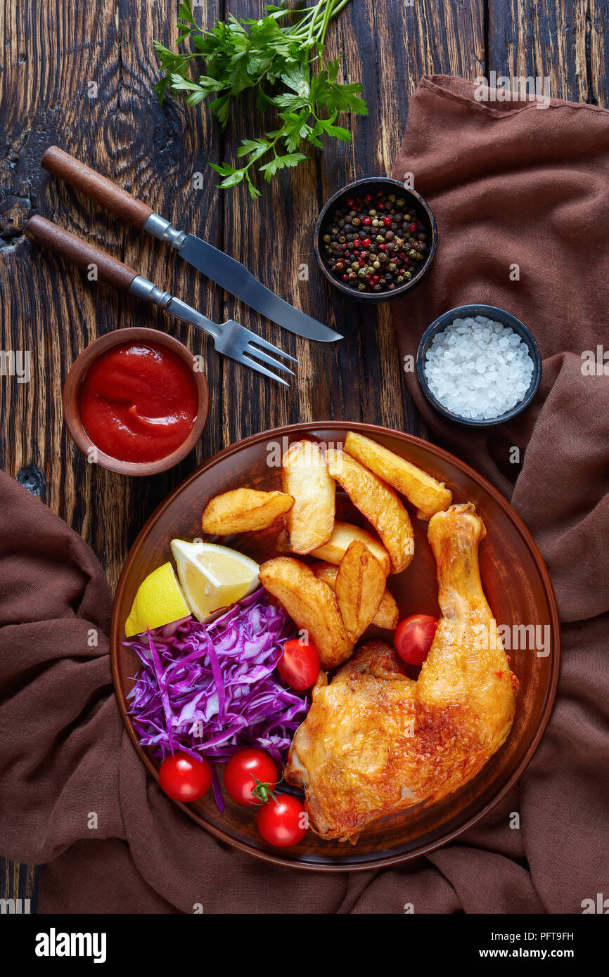 Patatas fritas ,pierna de pollo y ensalada coleslaw servido en un plato de barro rústico, con tenedor y cuchillo sobre una mesa de madera oscura, vista vertical de abov Foto de stock