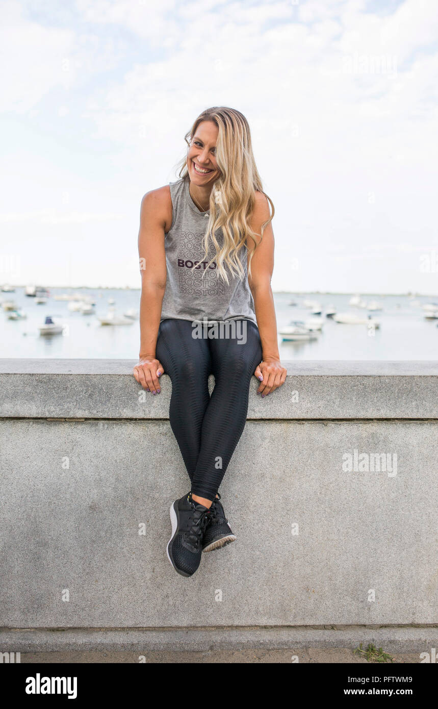 Sonriente joven mujer adulta en atuendo de Fitness sentado en muro de piedra con el océano en segundo plano. Foto de stock