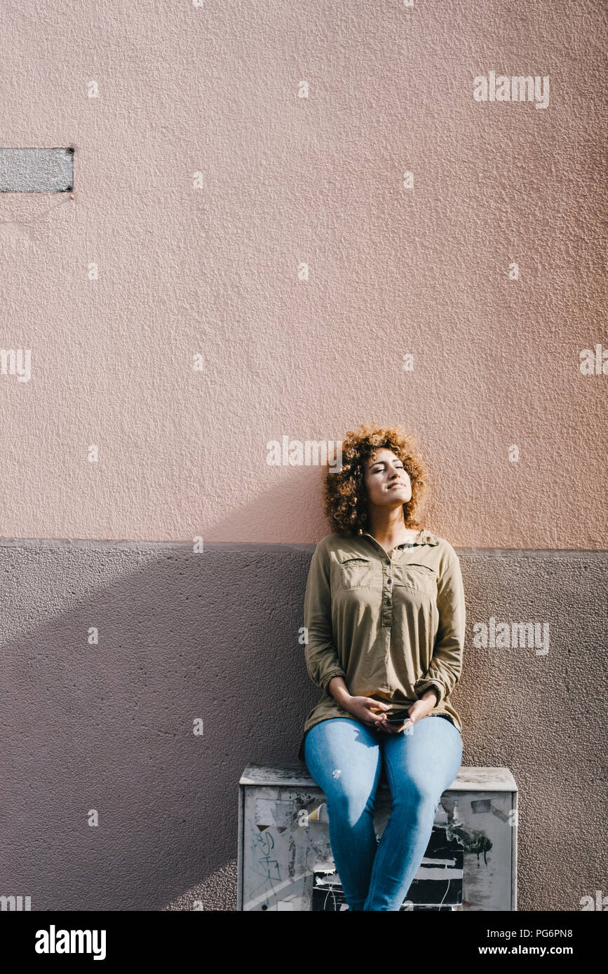 Mujer sentada delante de la pared de la casa, disfrutando del sol Foto de stock