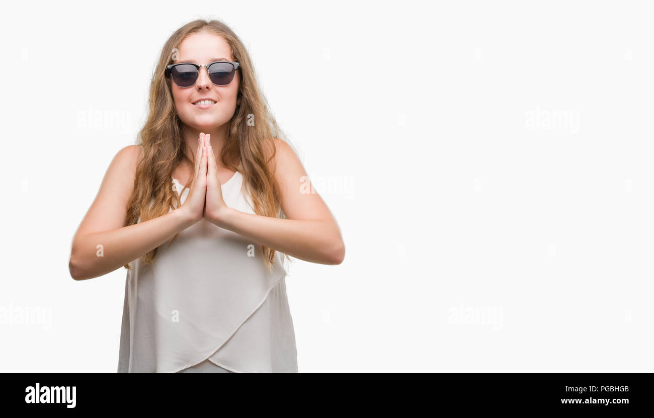 Joven Mujer rubia con gafas de sol rezando con las manos juntas pidiendo  perdón sonriendo confiada Fotografía de stock - Alamy