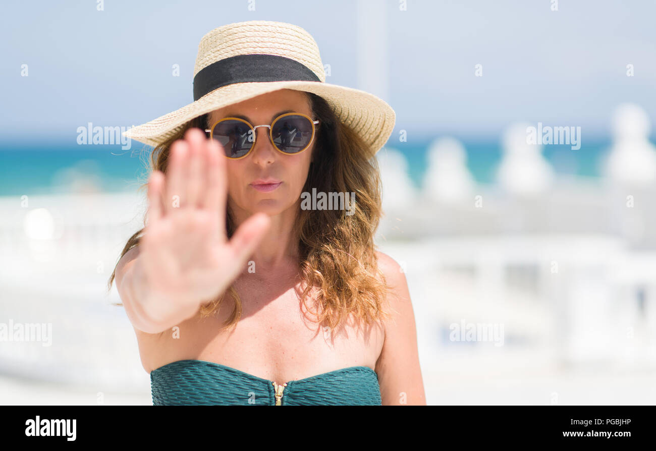 Mujer morena de mediana edad con gafas de sol junto a la piscina con la mano abierta haciendo la señal de stop, con graves y seguros de expresión, gesto de defensa Foto de stock
