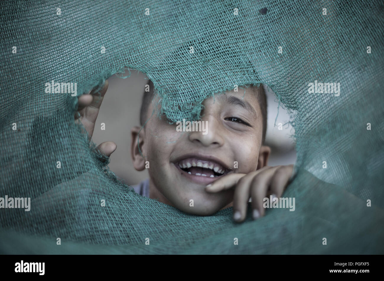 La ciudad de Gaza, en la Franja de Gaza, Gaza. 25 Aug, 2018. Un refugiado palestino kid visto sonreír a través de un orificio neto fuera de su hogar temporal en Beit Lahiya.Los Estados Unidos canceló más de doscientos millones de dólares de ayuda a los refugiados palestinos en la Franja de Gaza y Cisjordania. Crédito: Mahmoud Issa/SOPA Images/Zuma alambre/Alamy Live News Foto de stock