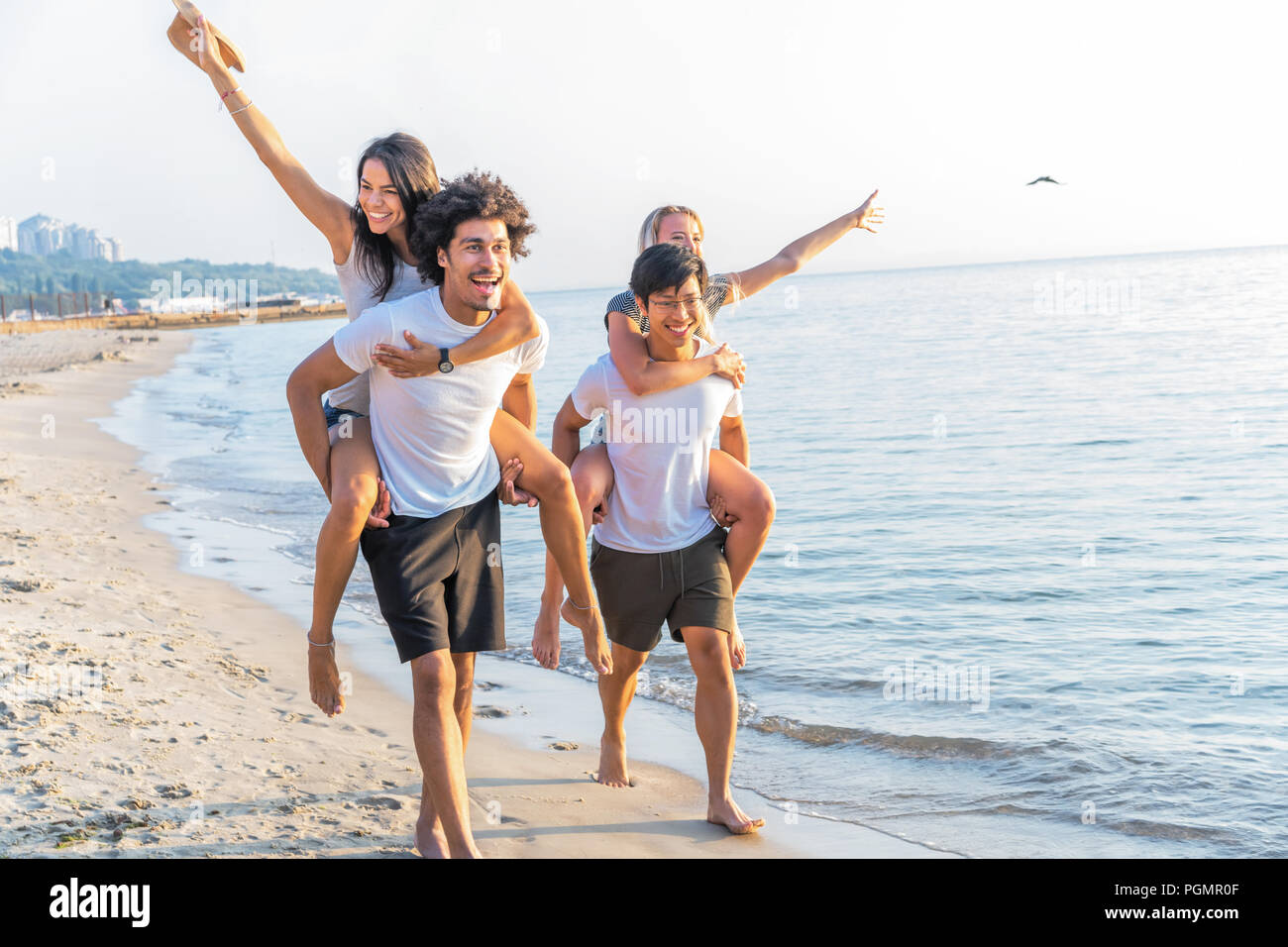 Grupo de amigos caminando por la playa, con los hombres que piggyback ride para novias. Feliz jóvenes amigos disfrutando de un día en la playa Foto de stock