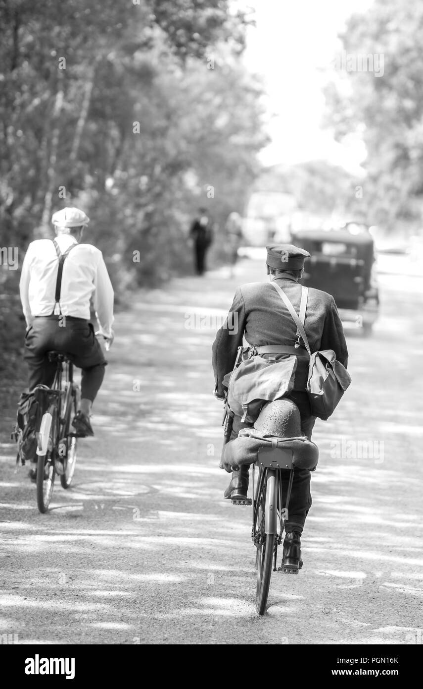 Vista nostálgica en blanco y negro, Crich Tramway Village, evento de verano de los años 40. Vista trasera de dos hombres montando bicicletas siguiendo vehículos antiguos bajo el sol. Foto de stock