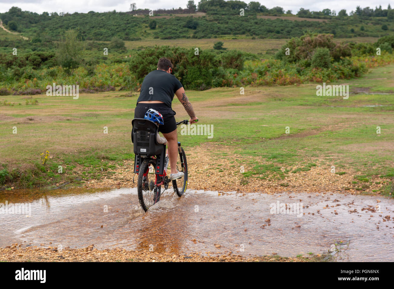 Hombre vestido con pantalones cortos en bicicleta a través del agua con un niño pequeño en la parte posterior de la bicicleta. Foto de stock