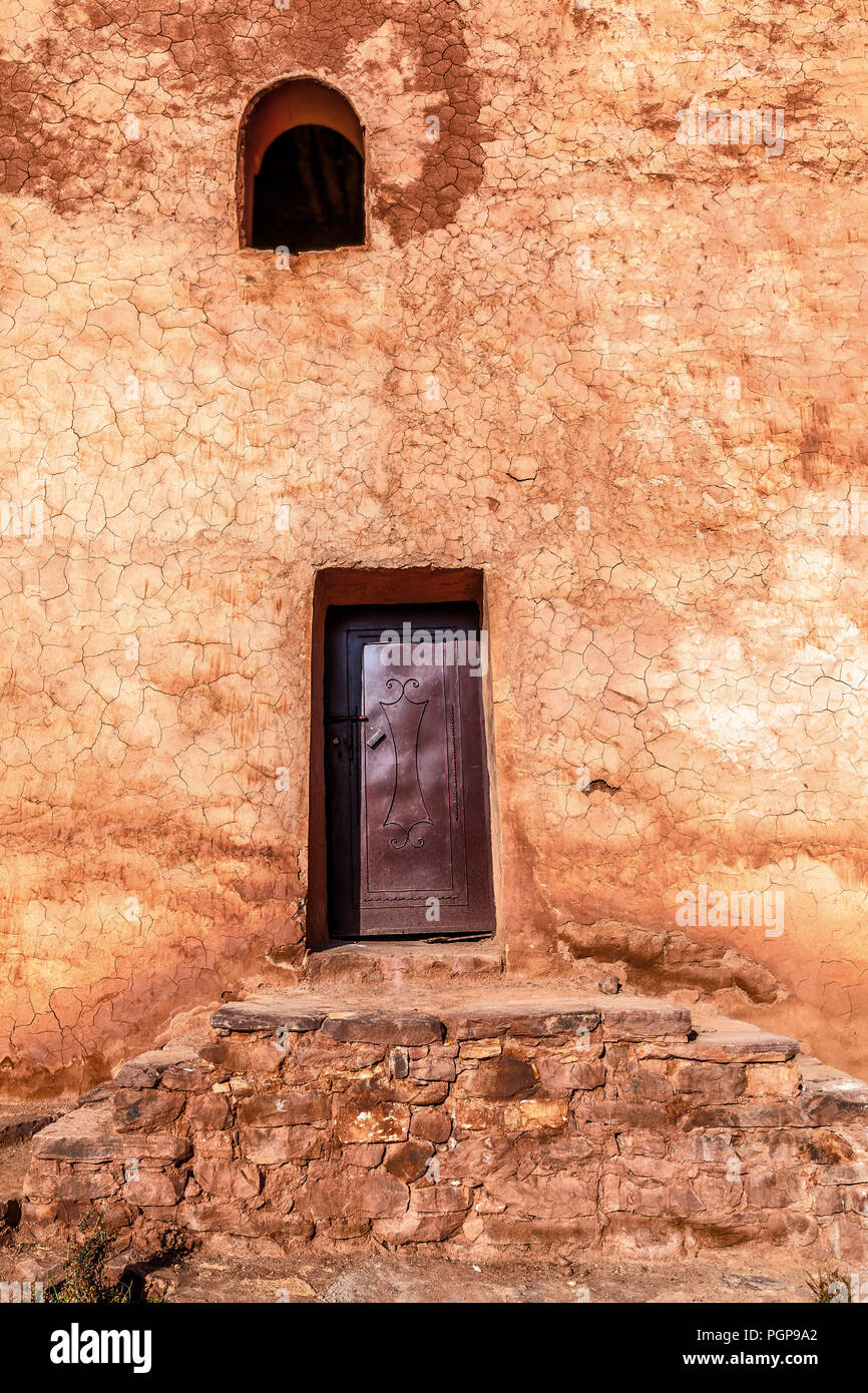 Marruecos pared de yeso rústico con escalones de piedra que conduce a una pequeña puerta de metal. Abrir ventana arqueada por encima. Ricos colores ocres. Pátina de edad. Vertical. Foto de stock