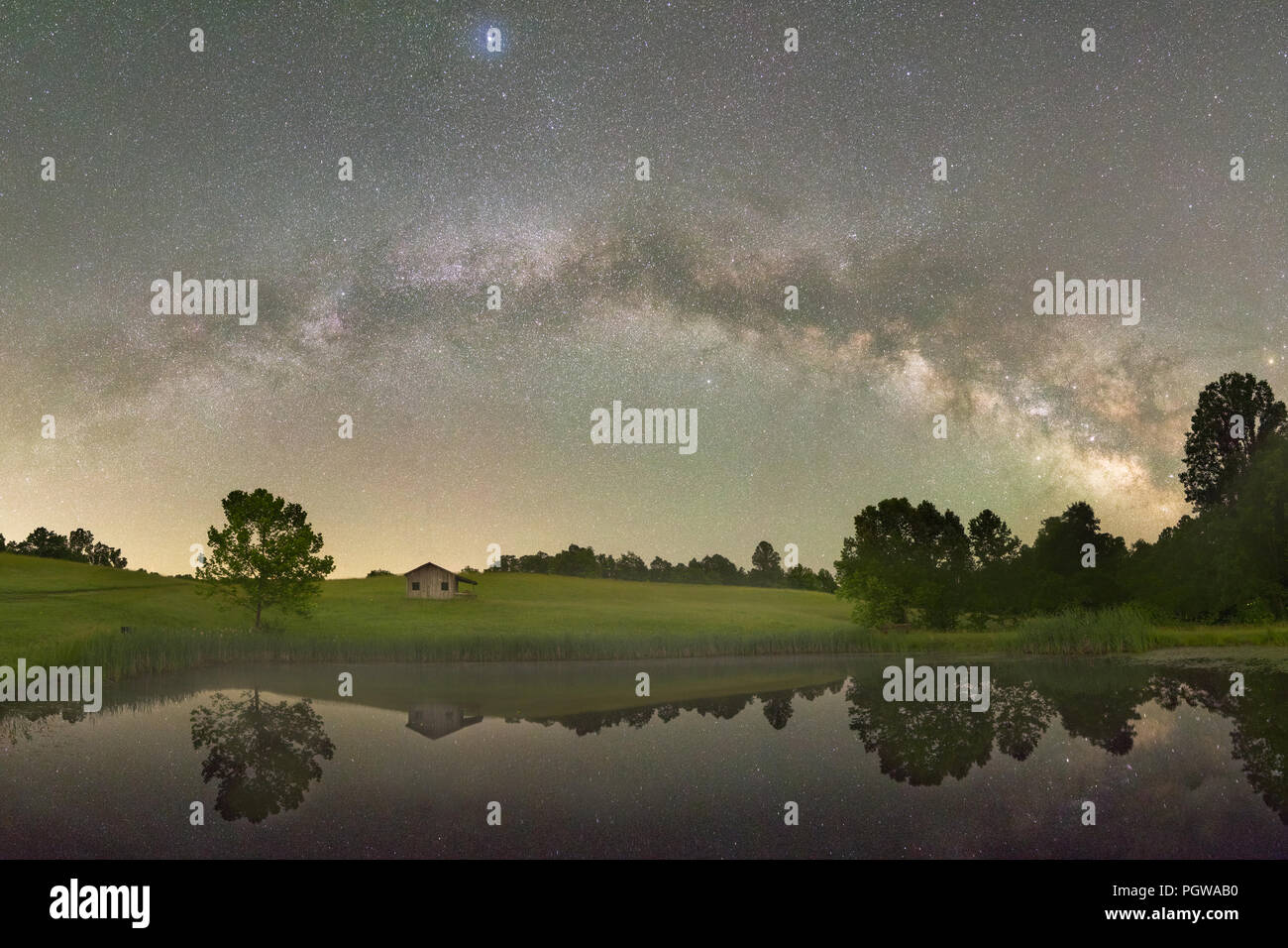 La longitud total de la Vía Láctea atraviesa la campiña de colinas y un viejo cobertizo, reflejado en un pequeño estanque por debajo de los cielos oscuros de West Virginia. Foto de stock