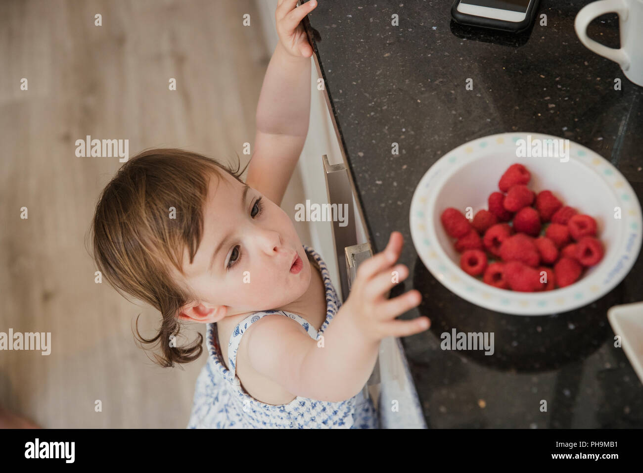 Un alto ángulo de vista de una niña en un kicthen standning y hasta llegar a través de la encimera de la cocina para coger algo de frambuesas. Foto de stock