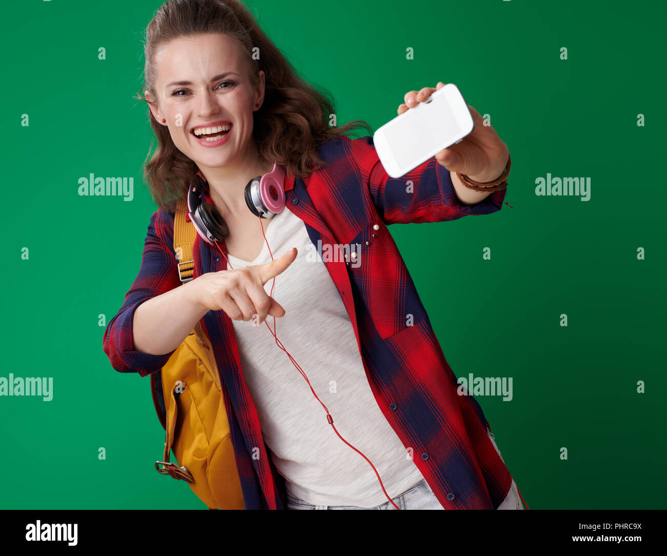 Sonriente joven estudiante mujer en una camiseta roja con mochila y los auriculares del smartphone apuntando contra el fondo verde Foto de stock
