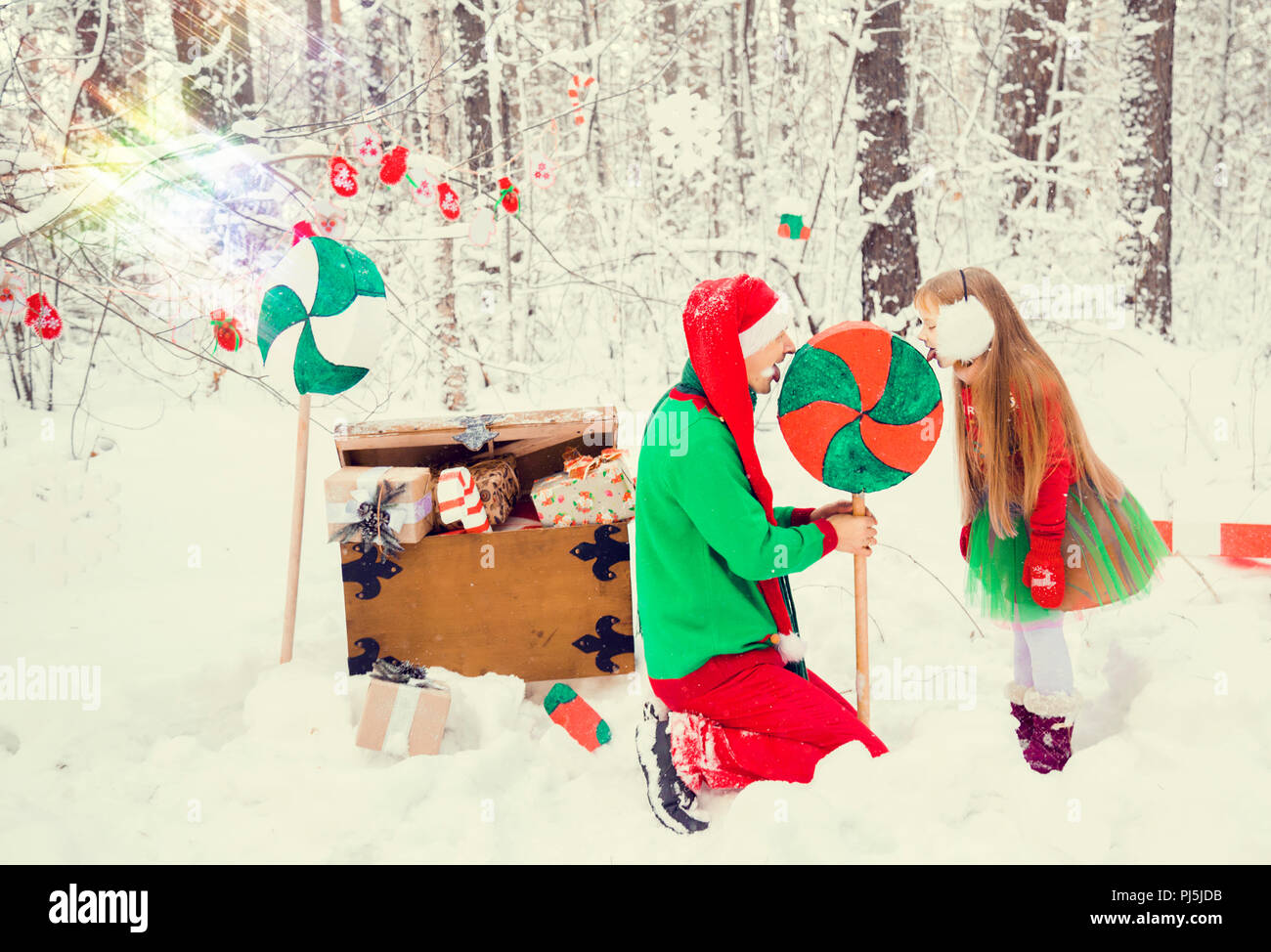 Padre e hija andando en trajes de flores tradicionales para los elfos ayudantes de Santa Claus en el bosque de invierno bajo la nieve con un cofre de regalos Foto de stock