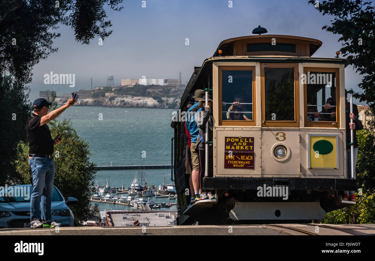 Cresterías Teleférico una colina en San Francisco mientras el hombre intenta coger un paseo con Alcatraz en el fondo Foto de stock