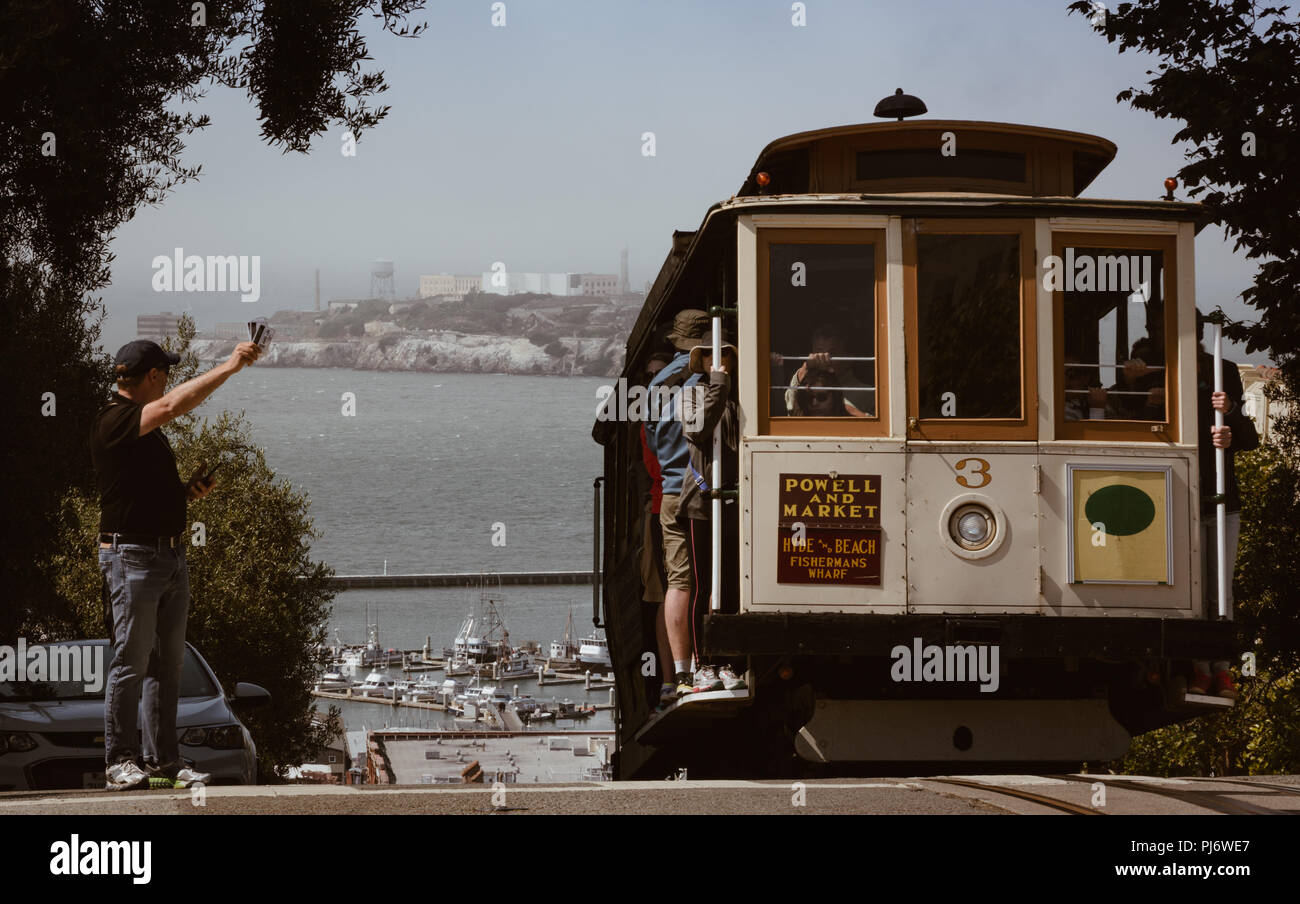 Cresterías Teleférico una colina en San Francisco mientras el hombre intenta coger un paseo con Alcatraz en el fondo Foto de stock