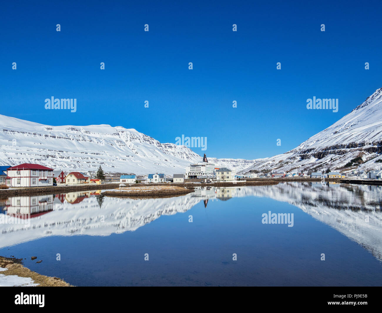 11 de abril de 2018; Seyðisfjörður Anuncios, Oriente Islandia - El pueblo se refleja en el fiordo, y el la línea de ferry Smyril MS Norrona. Foto de stock