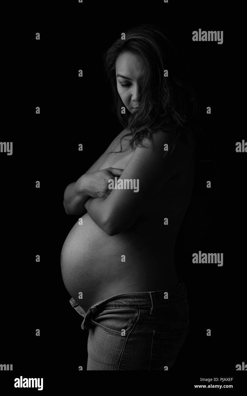Una embarazada mujer asiática posando al lado, mirando hacia abajo. Una artística oscuro retrato en blanco y negro. Foto de stock