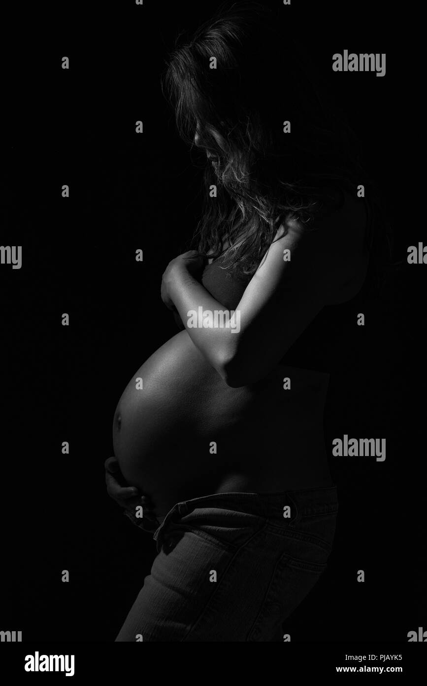 Una embarazada mujer asiática posando al lado, mirando hacia abajo. Una artística oscuro retrato en blanco y negro. Foto de stock