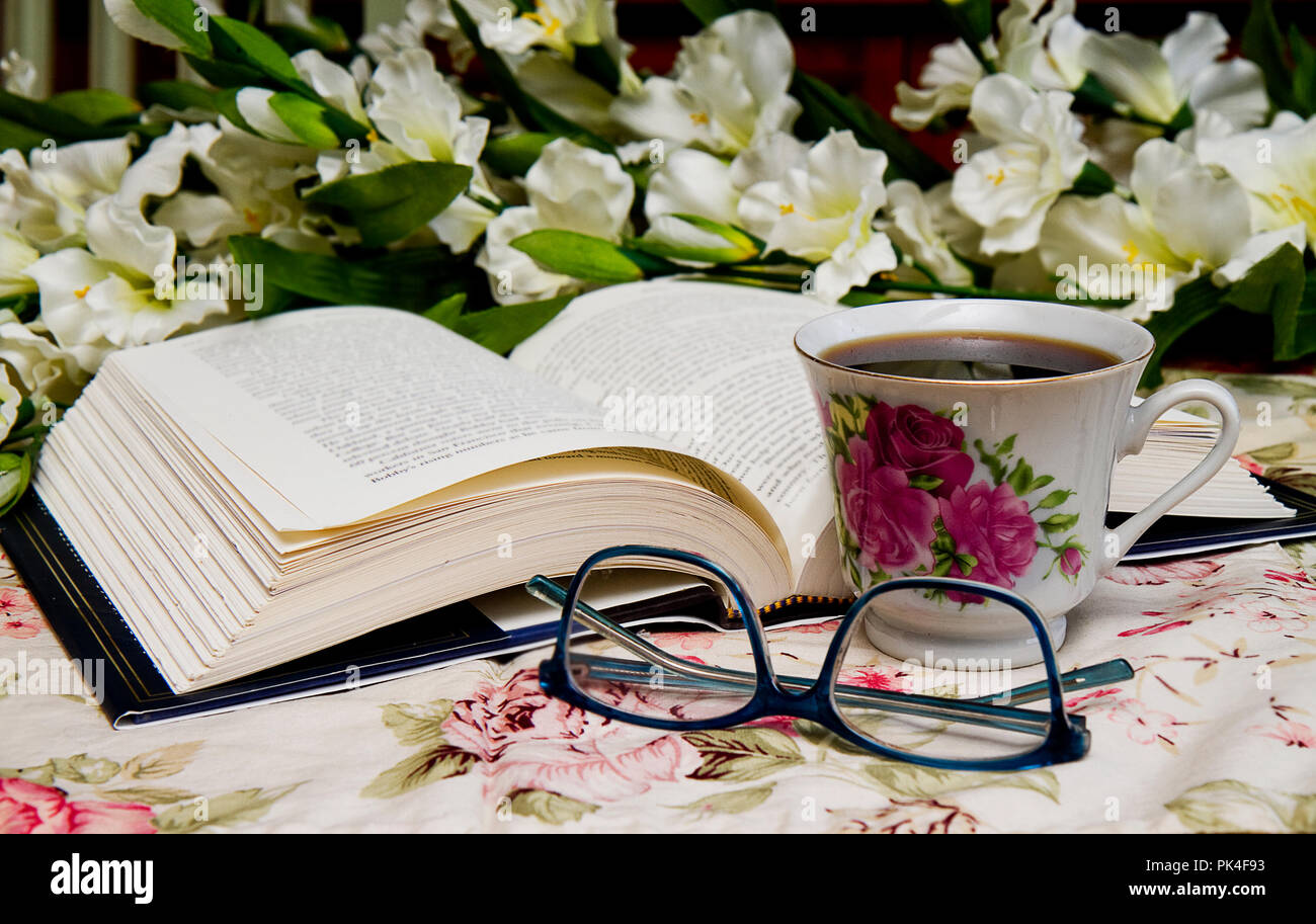 Libro abierto con un par de gafas y una taza de café sentado junto a él mientras lee. Ramo de flores en la imagen. Foto de stock