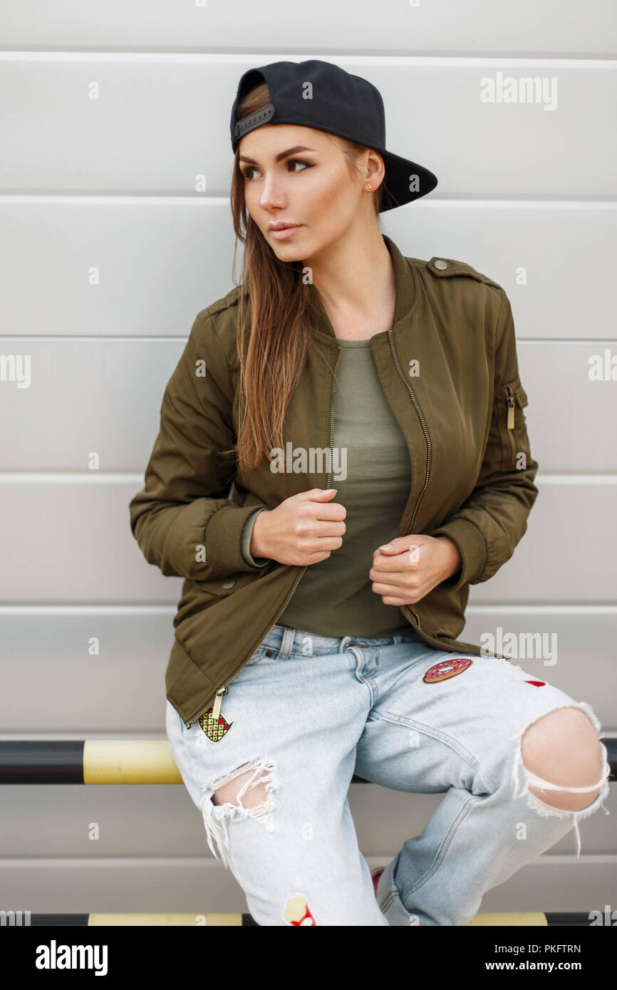 Hermosa muchacha elegante con pecas en una gorra de béisbol en un elegante chaqueta verde con pantalones vaqueros rasgados cerca de una pared de plata Foto de stock